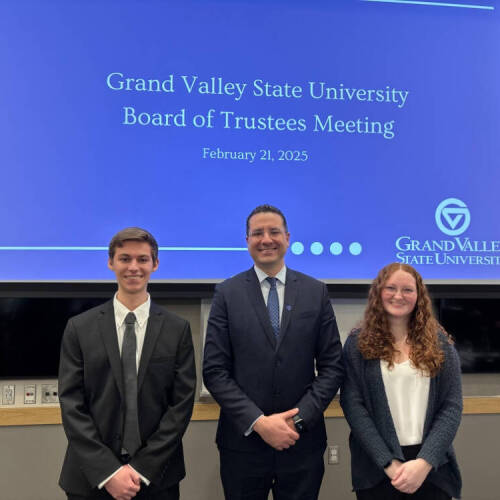 Dean Marouane Kessentini stands between two people in a room. Behind them is a blue drop banner with "Grand Valley State University Board of Trustees Meeting" wording.